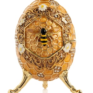 Könnte beinhalten: Verziertes, goldenes eiförmiges Schmuckkästchen mit Bienen- und Wabenmuster. Die Box hat einen Klappdeckel, dekorative Blumenakzente und wird von drei geschwungenen Beinen getragen. Das Ei ist mit Kristallen und einem facettierten Edelstein verziert.