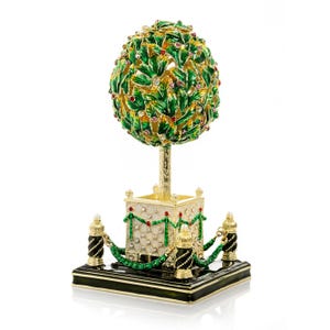 Könnte beinhalten: Eine dekorative, mit Juwelen besetzte Baumskulptur. Der Baum hat grüne Blätter mit goldenen Akzenten und ist mit roten und rosa Edelsteinen verziert. Er steht auf einem goldenen Ständer mit einer weißen, rautenförmigen Basis und ist von schwarzen und goldenen Pfosten umgeben, die durch eine grüne Kette verbunden sind.