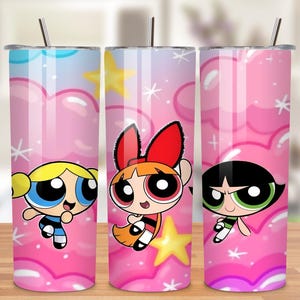 Puede incluir: Tres vasos de acero inoxidable con los personajes de dibujos animados de las Supernenas. Cada vaso tiene un diseño de personaje diferente sobre un fondo rosa con nubes, estrellas y copos de nieve. Los vasos tienen una pajita plateada.