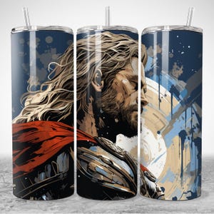 Puede incluir: Un vaso de acero inoxidable con una ilustraci&oacute;n estilizada de un guerrero con pelo largo y rubio, barba y capa roja. El dise&ntilde;o incluye un fondo azul con salpicaduras de pintura y un escudo. El vaso tiene una pajita transparente.