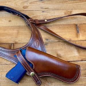May include: A brown leather shoulder holster with a matching belt and a blue handgun. The holster and belt are intricately stitched and feature decorative patterns. The leather has a rich, polished appearance, suggesting quality craftsmanship.