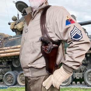 Op de afbeelding: Een man die een beige jas draagt met een leren schouderholster en een revolver. Hij staat voor een militaire tank met het woord "Fury" erop geschilderd. De tank heeft een groot nummer "2" aan de zijkant en een militair patch met een chevron op de mouw van de jas.