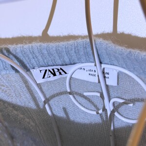 Puede incluir: Primer plano de un su&eacute;ter azul claro con una etiqueta ZARA. La etiqueta muestra la talla S y el origen NA. El su&eacute;ter est&aacute; hecho de un material suave y texturizado.
