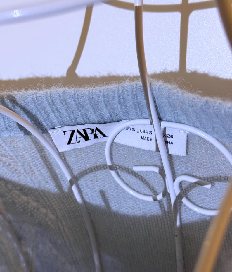 Puede incluir: Primer plano de un su&eacute;ter azul claro con una etiqueta ZARA. La etiqueta muestra la talla S y el origen NA. El su&eacute;ter est&aacute; hecho de un material suave y texturizado.