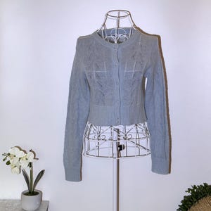 Puede incluir: Un c&aacute;rdigan corto azul claro con cuello redondo y cierre de botones. El su&eacute;ter presenta un punto trenzado en las mangas y un patr&oacute;n calado decorativo en la parte delantera. Se exhibe en un maniqu&iacute; de alambre blanco.