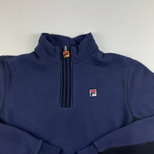 May include: Navy blue quarter-zip sweatshirt with a high collar. Features a black zipper with a red and orange logo at the top and a small logo on the chest. The sleeves have black accents.