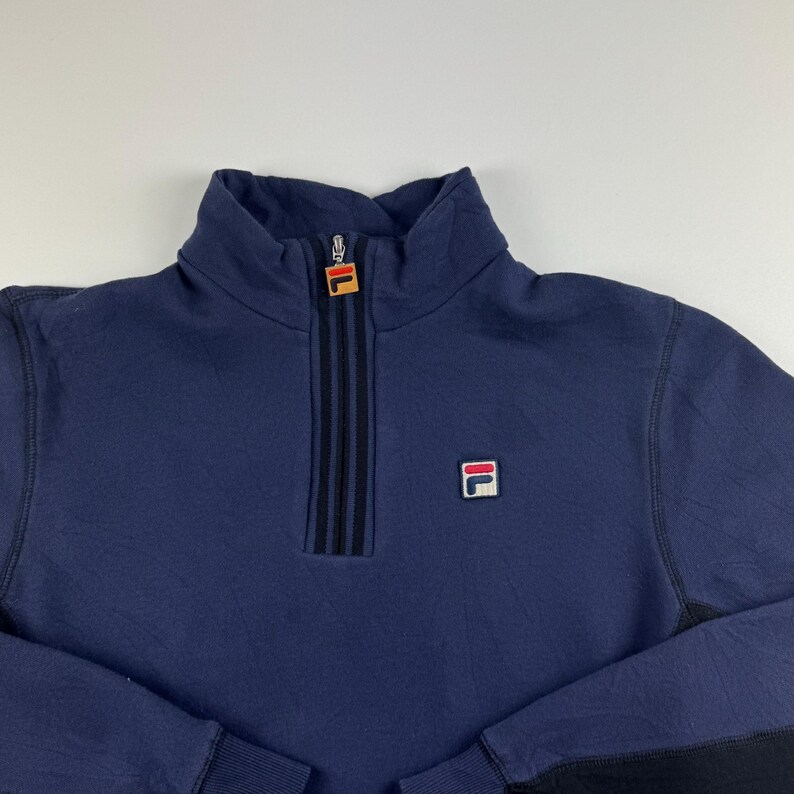 May include: Navy blue quarter-zip sweatshirt with a high collar. Features a black zipper with a red and orange logo at the top and a small logo on the chest. The sleeves have black accents.