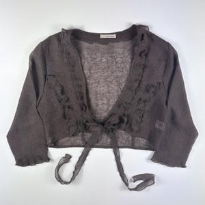 May include: A dark brown, open-front bolero with long sleeves and a delicate ruffle trim along the neckline and front edges. The bolero features a tie closure at the waist and is made of a sheer, lightweight fabric.