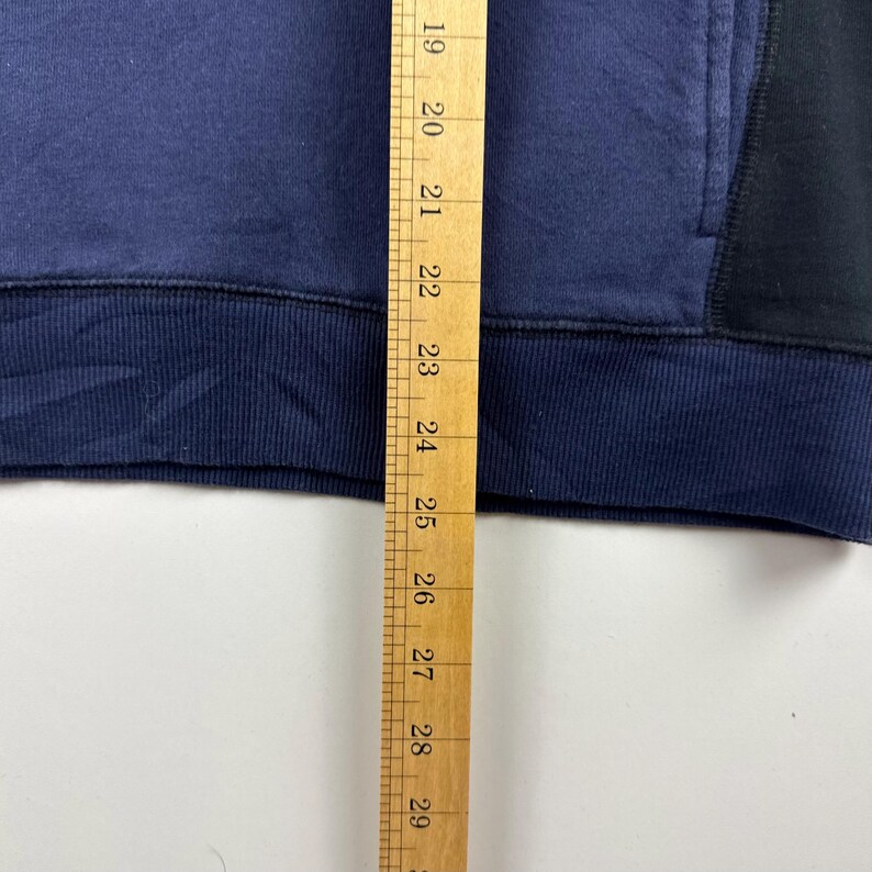 May include: A close-up of a navy blue and black sweatshirt. The sweatshirt has a color-blocked design with a navy blue body and black accents. A wooden ruler is placed vertically along the left side of the sweatshirt.