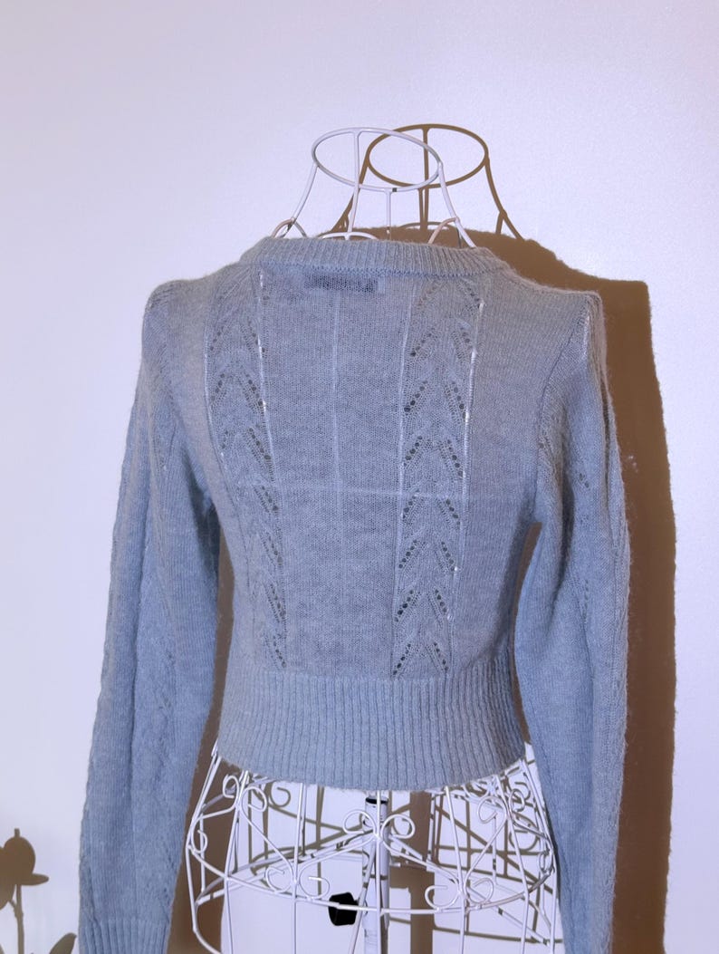 Puede incluir: Un su&eacute;ter azul claro con mangas largas y cuello redondo. La parte trasera del su&eacute;ter presenta un dise&ntilde;o calado decorativo con patrones verticales y diagonales. El su&eacute;ter se exhibe en un maniqu&iacute; de alambre blanco.