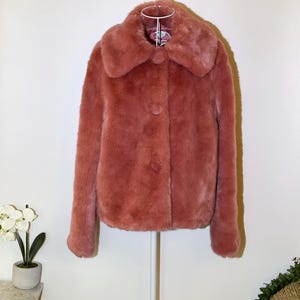 May include: A dusty rose faux fur coat with a large collar and three matching buttons. The coat is hanging on a white metal hanger against a white wall. The coat has long sleeves and a soft, plush texture.