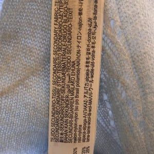 Puede incluir: Primer plano de una etiqueta de tela azul claro con texto en varios idiomas. La etiqueta indica la composici&oacute;n de la tela: 50% nailon, 25% alpaca y 25% lana. La etiqueta est&aacute; adherida a una tela azul claro y transparente.