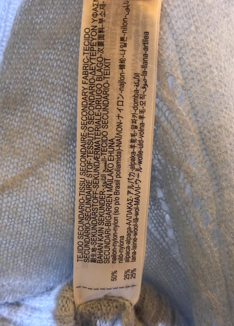 Puede incluir: Primer plano de una etiqueta de tela azul claro con texto en varios idiomas. La etiqueta indica la composici&oacute;n de la tela: 50% nailon, 25% alpaca y 25% lana. La etiqueta est&aacute; adherida a una tela azul claro y transparente.