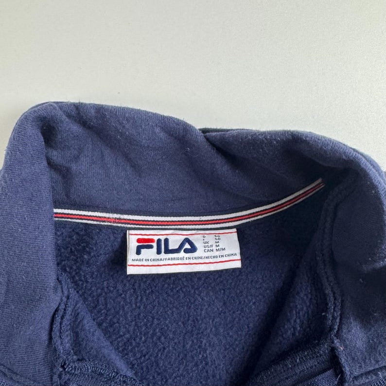 May include: Close-up of a navy blue FILA sweatshirt collar. The collar has a red and white stripe detail. The FILA logo is visible on a white tag, along with text indicating the item was made in China.