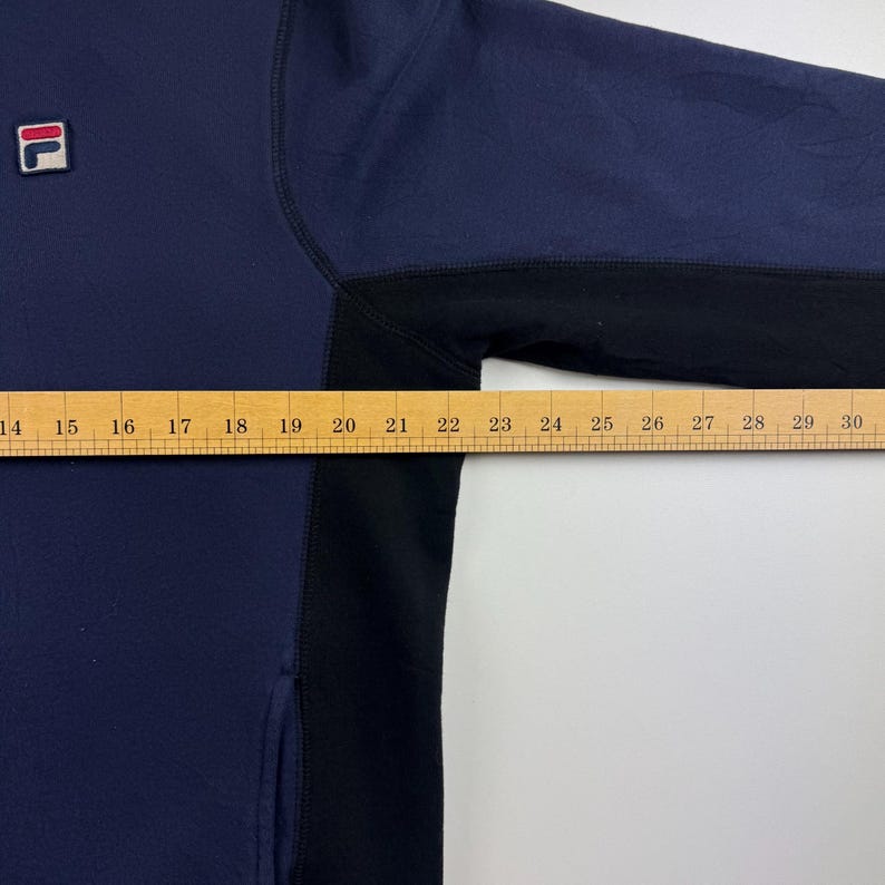 May include: A navy blue and black sweatshirt with a small logo on the upper left chest. The sweatshirt has a color block design and a measurement ruler is placed across the garment. The ruler measures approximately 16 inches.