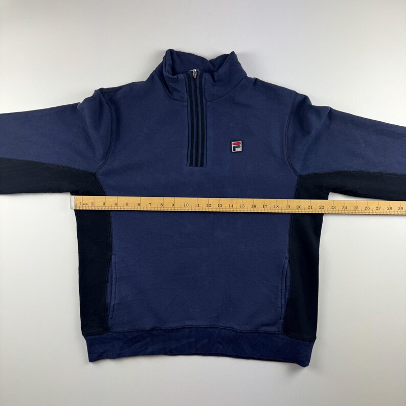 May include: Navy blue sweatshirt with a quarter-zip closure and black side panels. The Fila logo is on the chest. The sweatshirt has a high collar and long sleeves.