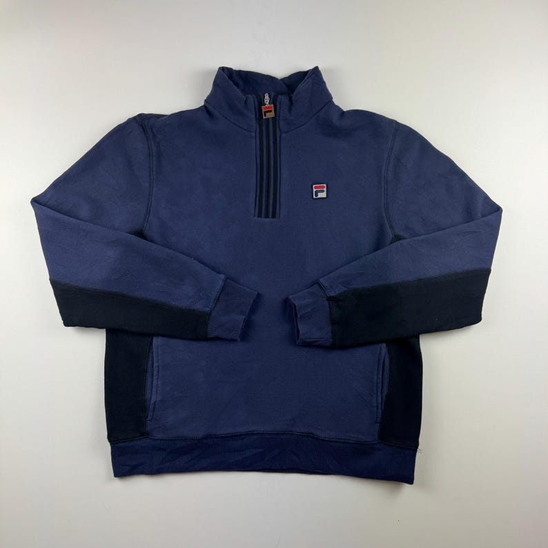 May include: Navy blue sweatshirt with a quarter-zip closure and a small logo on the chest. The sleeves have black panels. The collar and zipper pull feature red accents. A casual, sporty top.