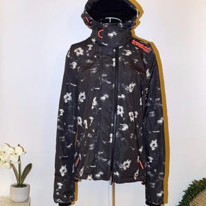 Peut inclure: Veste à capuche noire ornée d'un motif floral. La veste est dotée d'une fermeture éclair intégrale, d'un col montant et du mot "Superdry" en orange sur la poitrine droite. Elle présente des poignets noirs et des touches d'orange.