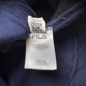 May include: Close-up of a navy blue garment tag with the brand name "FILA" in white. The tag includes a QR code and text detailing the fabric composition and origin. The garment is made in China.