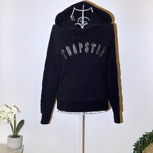 May include: Black hoodie with the word "TRAPSTAR" in white, arched across the chest. The hoodie has a hood, a front pocket, and white stripes down the sleeves. The hoodie is displayed on a white mannequin.