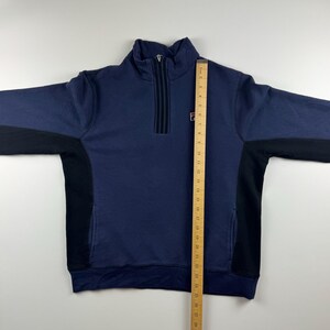 May include: A navy blue sweatshirt with a quarter-zip closure and black side panels. The sweatshirt features a small logo on the chest and a high collar. A ruler is placed next to the sweatshirt to measure its length.