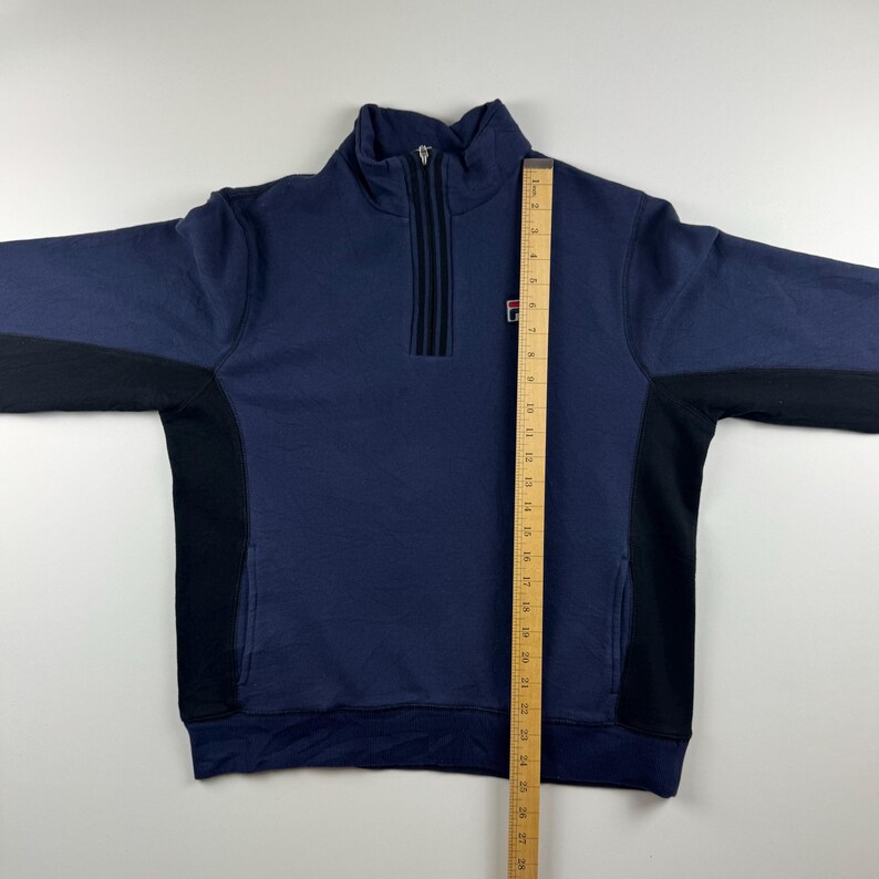 May include: A navy blue sweatshirt with a quarter-zip closure and black side panels. The sweatshirt features a small logo on the chest and a high collar. A ruler is placed next to the sweatshirt to measure its length.