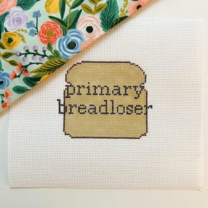 May include: A cross-stitched canvas with the words "primary breadloser" in black thread, set against a tan bread-shaped background. A floral fabric with pink, blue, and yellow flowers and green leaves is in the upper left corner.