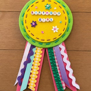 May include: A circular felt badge featuring a yellow center with the text BIRTHDAY GIRL spelled out in white letter beads. The badge is decorated with small flower appliques and sequins, surrounded by a lime green felt border and colorful ribbons.