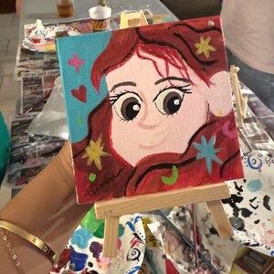 May include: A small square painting on a wooden easel. The painting features a cartoon face with large eyes, red hair, and colorful star and heart accents against a blue background. The artist's signature is visible.