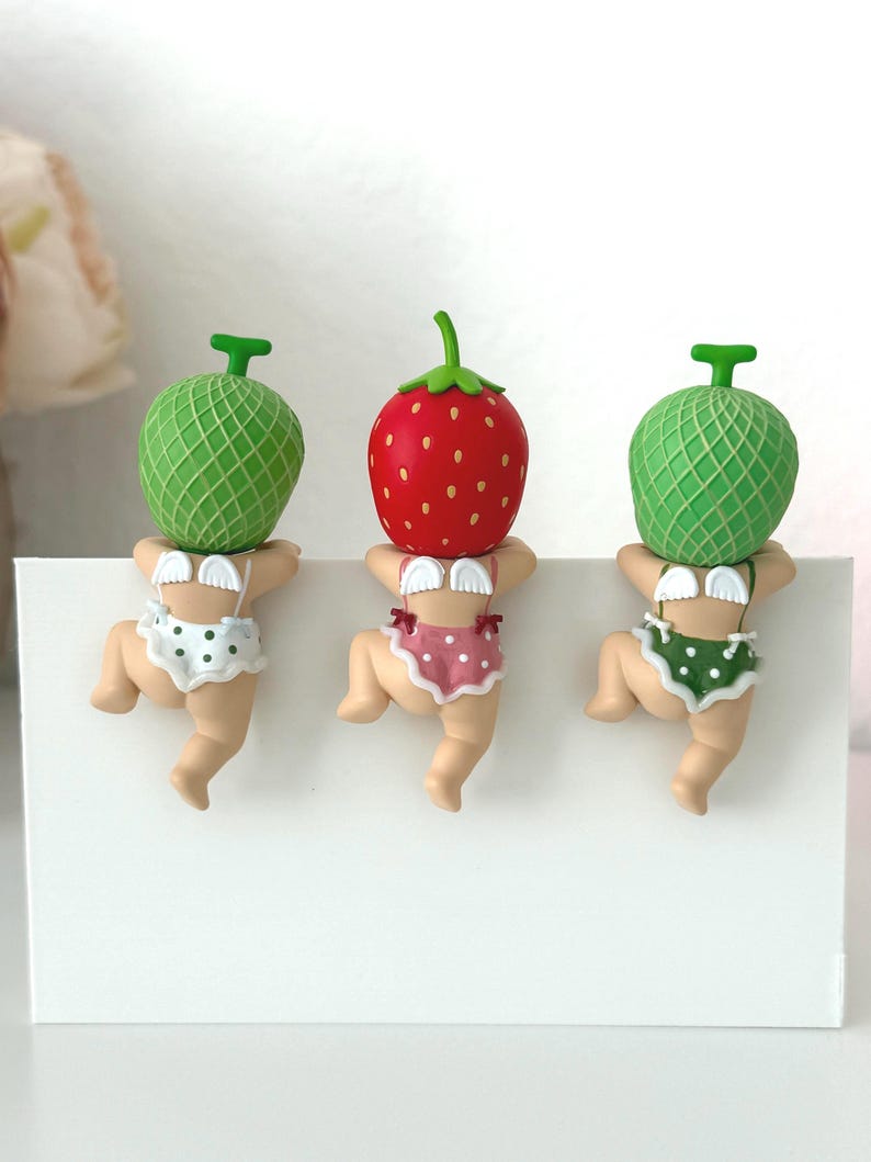 May include: Three small figurines featuring baby-like characters with fruit heads. Two have green melon heads and one has a red strawberry head. Each character wears white angel wings and polka-dot ruffled bottoms while climbing a white edge.
