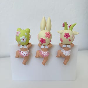 May include: Three small figurines of babies with animal-like heads, including a frog, a rabbit, and a goat. Each figure features a pale skin tone, small white wings on the back, and patterned underwear. The heads are decorated with pink cherry blossom motifs.