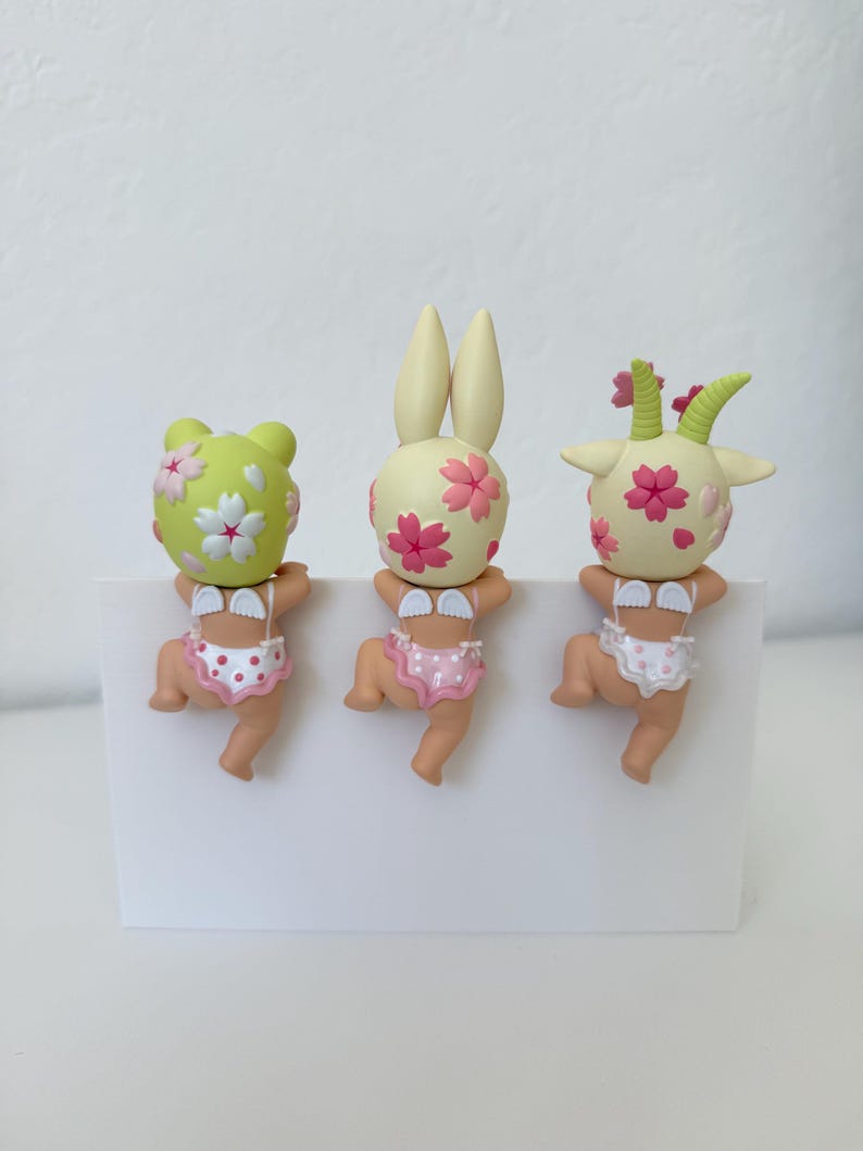 May include: Three small figurines of babies with animal-like heads, including a frog, a rabbit, and a goat. Each figure features a pale skin tone, small white wings on the back, and patterned underwear. The heads are decorated with pink cherry blossom motifs.