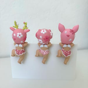 May include: Three small figurines featuring pink heads with white cherry blossom patterns, small white wings, and pink polka-dot bottoms. The left figure has green antlers, the center has rounded ears, and the right has pointed ears, all hanging.