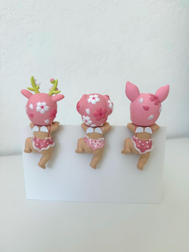 May include: Three small figurines featuring pink heads with white cherry blossom patterns, small white wings, and pink polka-dot bottoms. The left figure has green antlers, the center has rounded ears, and the right has pointed ears, all hanging.