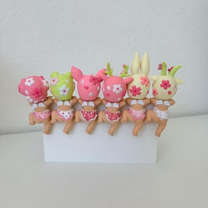 May include: A row of six small, stylized figurines with animal-like features, each wearing polka-dot underwear and small white wings. The figures have heads decorated with pink cherry blossom patterns in various colors including pink, green, and cream.
