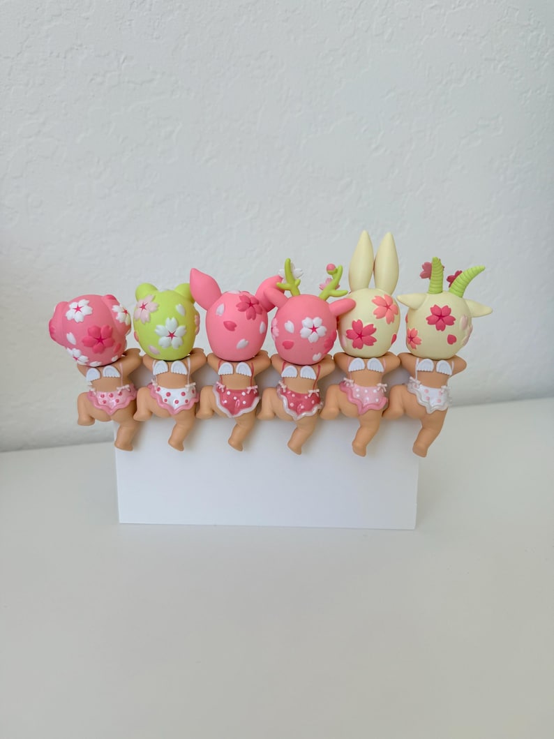 May include: A row of six small, stylized figurines with animal-like features, each wearing polka-dot underwear and small white wings. The figures have heads decorated with pink cherry blossom patterns in various colors including pink, green, and cream.