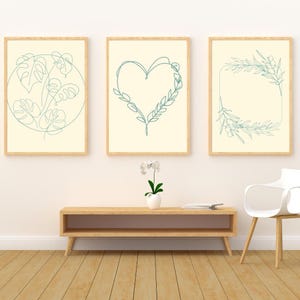 以下が含まれることがあります： 3つの額入りアートプリント。モンステラの葉のデザイン、葉の付いたハート、オリーブの枝の長方形のフレームが特徴です。アートワークは、木製のフレームが付いた、クリーム色の背景に水色の線で描かれています。