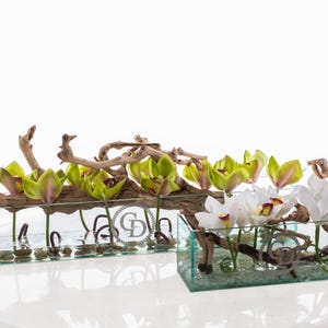 May include: Two rectangular glass vases with floral arrangements. One vase features green and pink orchids, the other white orchids. Both arrangements include driftwood and are set on a white surface.