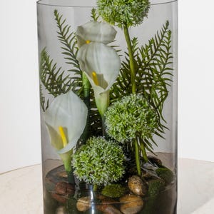 May include: A clear glass cylinder vase filled with white calla lilies, green fern-like leaves, and spherical green plants. The base contains small stones and moss, creating a natural, decorative arrangement.