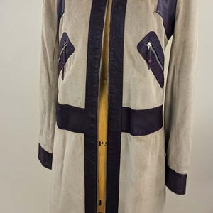 May include: A long, beige suede coat featuring dark purple leather accents on the collar, front placket, waist, cuffs, and shoulder panels. The coat includes a full-length front zipper and two zippered chest pockets with leather trim.