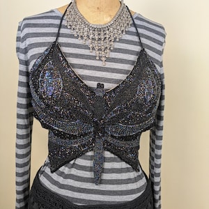 Op de afbeelding: Een zwarte, met kralen bezette vlindertop met iriserende accenten. De top is in halterstijl en heeft een gedetailleerd vlinderontwerp. Een zilveren ketting wordt bij de top gedragen. De top wordt op een mannequin getoond.