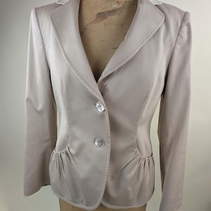 May include: A light beige blazer featuring a notched lapel, long sleeves, and a two-button front closure. The garment includes two front pockets with gathered, pleated detailing at the base, creating a structured silhouette.