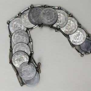 Könnte beinhalten: Ein silberfarbenes Armband mit mehreren runden Münzen, die miteinander verbunden sind. Jede Münze zeigt ein detailliertes Design und die Zahl "20". Das Armband hat einen Verschluss und wird vor einem neutralen Hintergrund präsentiert.