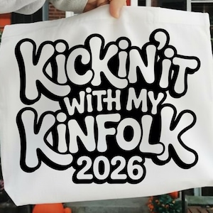 May include: A white tote bag with the phrase "Kickin' it with my Kinfolk 2026" in a bold, black, bubble-style font. The bag is held up by a person, and the background is slightly blurred.