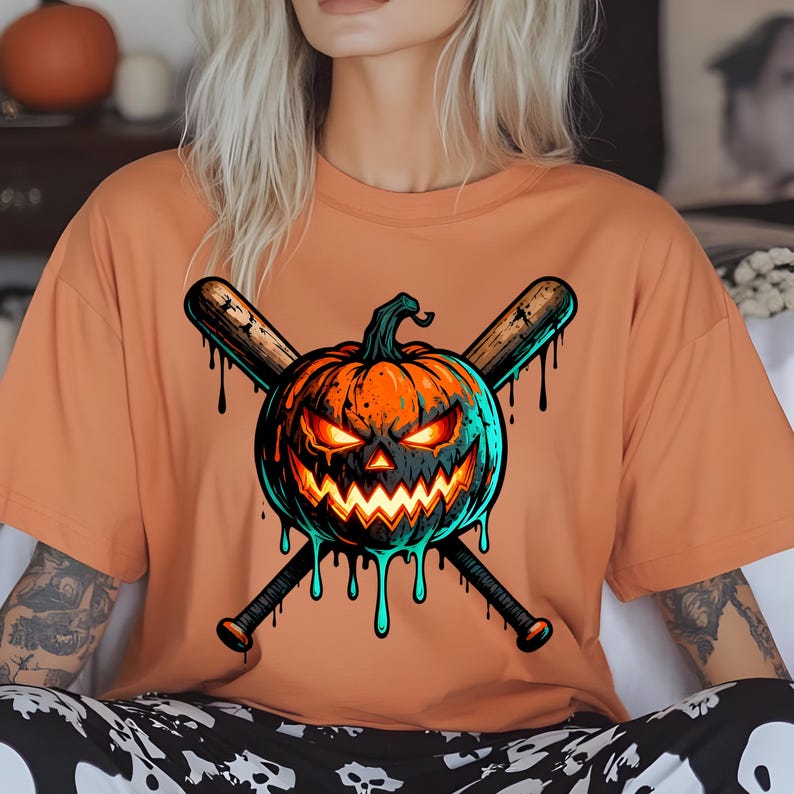 May include: Orange t-shirt featuring a Halloween-themed graphic. The design includes a jack-o'-lantern with glowing orange eyes and a jagged mouth, crossed baseball bats, and dripping teal accents. The shirt is a solid orange color.