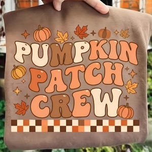 May include: A brown sweatshirt with the words "PUMPKIN PATCH CREW" in a retro font. The design includes pumpkins, leaves, and stars in shades of orange, brown, and cream. A checkered border is at the bottom.