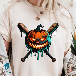 May include: Cream-colored t-shirt featuring a Halloween-themed graphic. The design includes a jack-o'-lantern with glowing orange eyes and a menacing grin, crossed with two wooden baseball bats. The pumpkin has teal accents and dripping teal paint.
