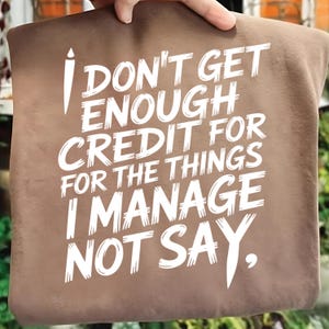 May include: A brown tote bag with the white text: "I DON'T GET ENOUGH CREDIT FOR THE THINGS I MANAGE NOT SAY." The bag is held up by a hand. The tote bag is a neutral color.
