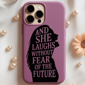 May include: A mauve phone case featuring a black silhouette of a woman's profile with long hair. The text "AND SHE LAUGHS WITHOUT FEAR OF THE FUTURE" is printed in black. The case has a camera cutout and is surrounded by pearls and flowers.