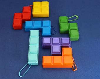 May include: A collection of colorful, plastic Tetris-style puzzle pieces in red, yellow, blue, green, purple, and orange. Each piece has a small chain attached. The pieces are arranged on a dark blue surface.